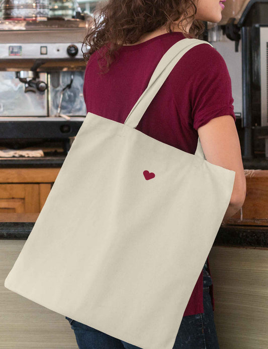 Frau trägt einen Baumwollbeutel naturfarben, mit Bodenfalte, langen Henkeln zum besseren Tragen und einem kleinen weichen Herz aus Flock