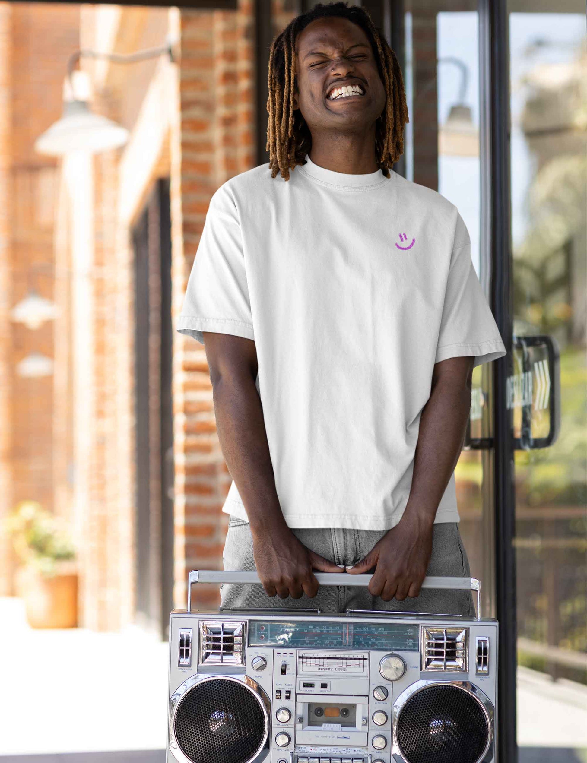Lachender Mann mit lockerem oversized T-Shirt in schlichtem Weiß und kleinem gespraytem pinken Smiley vorne